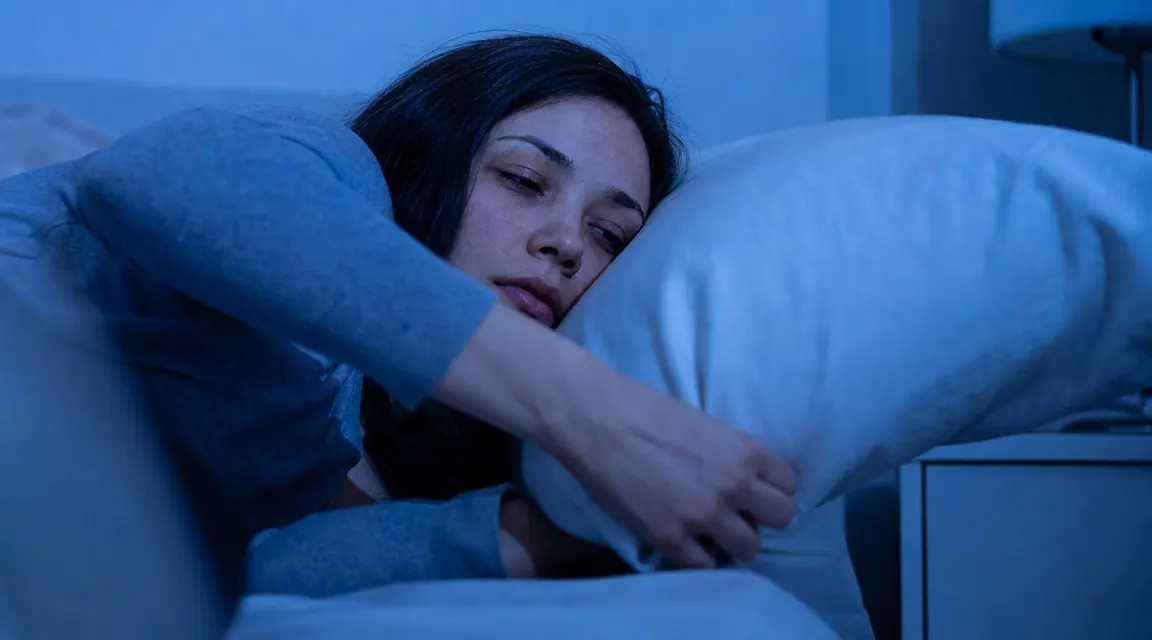 Person reaching over to flip their pillow at night, frustrated look, dim bedroom light