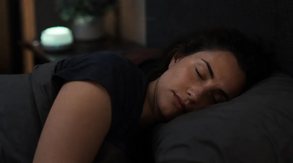 Person sleeping peacefully in a dark bedroom while a small sound machine glows on the nightstand